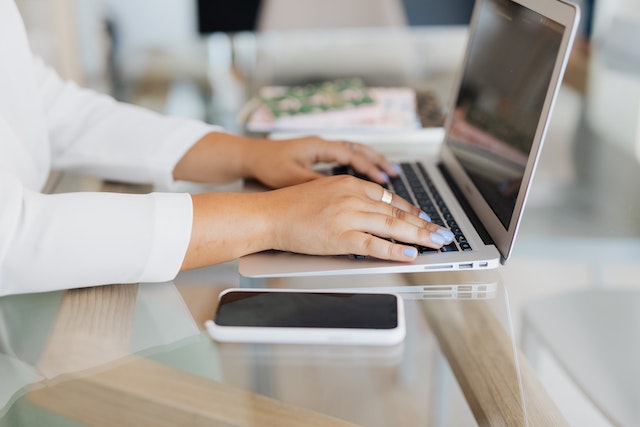 typing on a laptop with a cell phone next to them on a grey desk